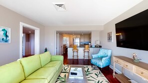 A living room with a green sofa, a blue patterned chair, a coffee table with remote controls, a kitchen in the background, and a TV mounted on the wall.