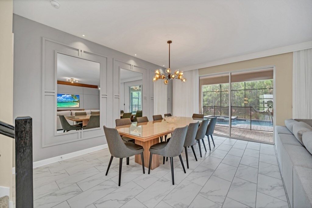 Uma sala de jantar moderna com uma mesa grande e cadeiras, um candelabro e vista para uma piscina e vegetação lá fora.