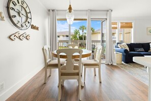A dining area with a round table and chairs, a wall clock, and a view of a balcony with a railing and palm trees. - Hoosier Holiday 1 Bedroom Condo by RedAwning (Destin)