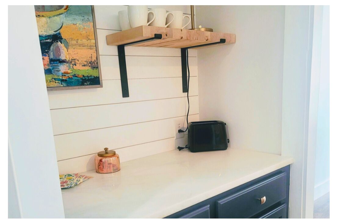 Eine Küche mit weißer Arbeitsplatte, einem Toaster, einer kleinen Schale und einem Regal mit Tassen und einem Bild.