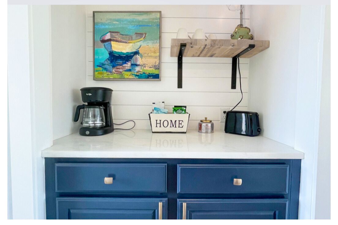 A kitchen counter with a coffee maker, a "HOME" sign, and a shelf with cups and a decorative item.