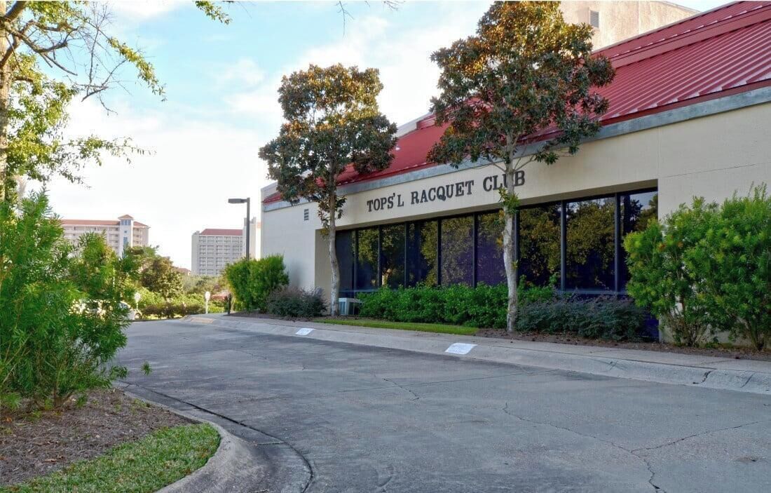 En bygning med et skilt, der viser 'TOPS'L RACQUET CLUB', og en parkeringsplads foran.