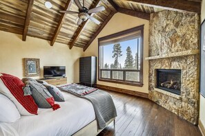 Quarto com cama grande, televisão, lareira de pedra e janela com vista para árvores.
