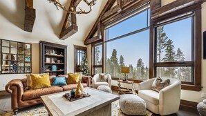 A spacious living room with a large window offering a view of trees, a leather sofa with colorful cushions, a wooden coffee table, and a rustic chandelier. - The Great Escape: Breckenridge Home with Hot Tub and Mountain Views (Breckenridge)