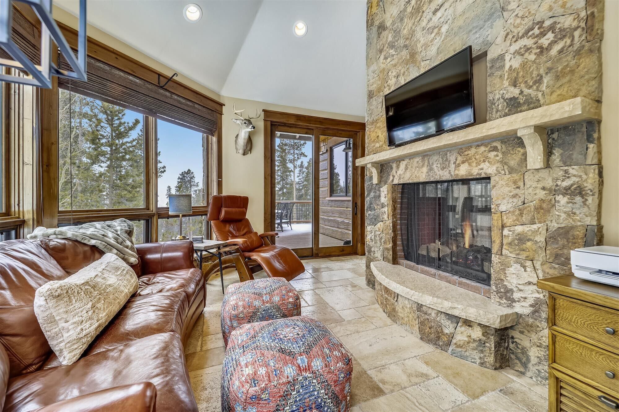 A cozy living room with a stone fireplace, leather furniture, and large windows offering a view of the outdoors.