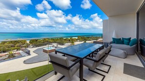 Ein Balkon mit Glastisch und Stühlen, ein Sofa mit Türkis-Kissen und Blick aufs Meer.