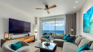 Ein modernes Wohnzimmer mit Flachbildfernseher, bequemen Sitzmöglichkeiten, einem Deckenventilator und einem großen Fenster mit Meerblick.