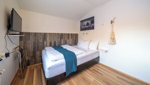 A modern bedroom with a wooden headboard, a bed with white linens and a teal throw, a mounted TV, and a wall-mounted towel rack. - Guesthouse Mikael (Höfn)