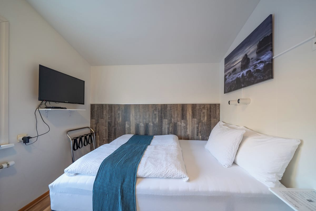 A hotel room with a bed, a flat-screen TV mounted on the wall, a wooden headboard, and a painting of a coastal scene.