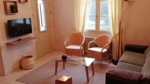 A living room with a television, two wicker chairs, a glass coffee table, a sofa, and a window with curtains.