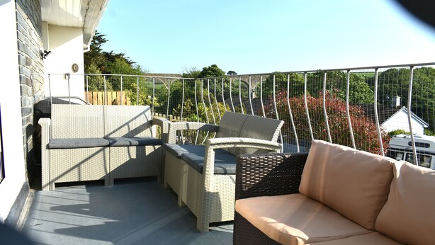 Ein Balkon mit Korbmöbeln, darunter eine Sitzecke mit Sofa und Stühlen, und Blick auf Grünflächen und Gebäude im Hintergrund.