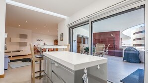 Une cuisine moderne dotée d’un îlot central, d’un coin repas avec des chaises et d’une vue sur une terrasse extérieure, accessible par une porte vitrée.