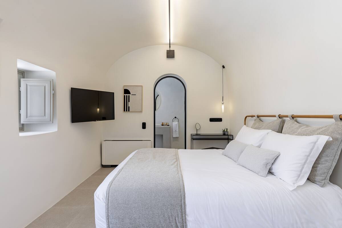 Ein modernes Hotelzimmer mit einem großen Bett, einem flachbildfernseher an der Wand, einem kleinen Nachttisch mit Lampe und einer Tür zu einem Badezimmer.