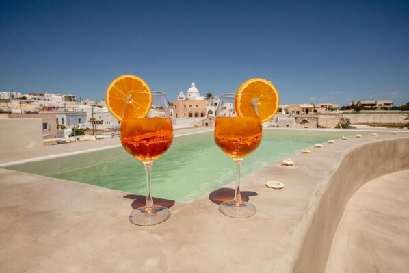 Two glasses of amber-colored beverage with orange slices on a rooftop terrace with a pool and buildings in the background. - Utopia Suites Santorini (Santorini)