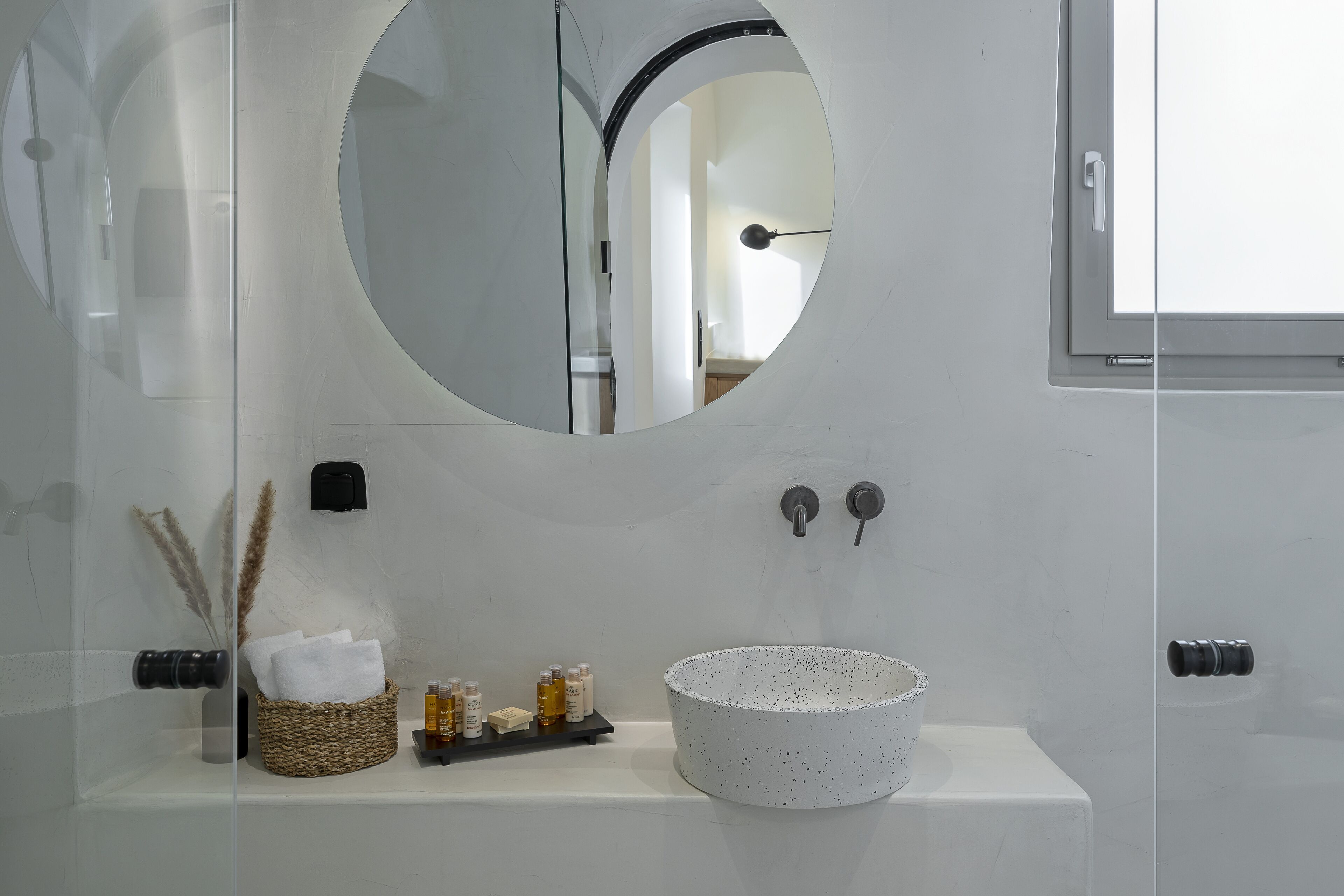 Un baño con un espejo redondo, un lavamanos blanco con un lavabo moteado, un estante con artículos de aseo y un perchero para toallas.