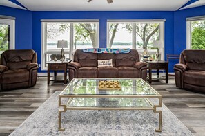A living room with a brown leather sofa, a glass coffee table, and a view of trees through large windows.