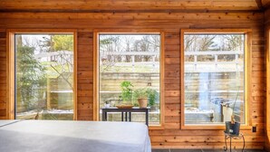 A wooden cabin with three large windows, a bed, a small table with plants, and a heater. - Hiking, Hot Tub, Lake Front, Dock, Swimming (Canandaigua)