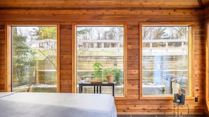 A wooden cabin with three large windows, a bed, a small table with plants, and a heater. - Hiking, Hot Tub, Lake Front, Dock, Swimming (Canandaigua)