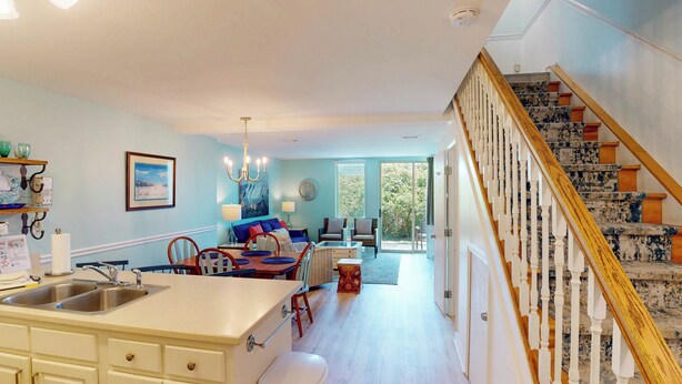 A spacious kitchen with a central island, a dining area with a blue sofa, and a staircase with a wooden railing. - Villa 389 2 bedroom, 1.5 bathroom (Jekyll Island)