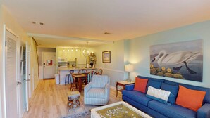 A living room with a blue sofa, a coffee table, and a painting of swans. There is a dining area with a table and chairs, and a kitchen in the background. - Villa 389 2 bedroom, 1.5 bathroom (Jekyll Island)