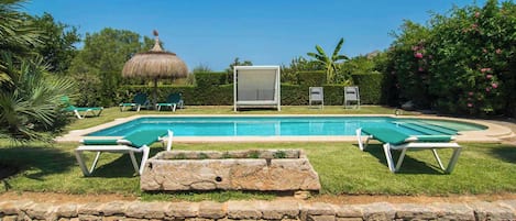 Una piscina con sillas de descanso, una estructura de techo de paja y abundante vegetación.