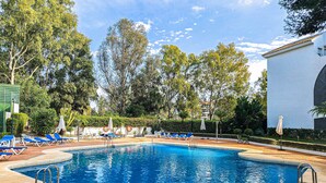 A swimming pool with blue water, surrounded by lounge chairs and umbrellas, with trees and a building in the background. - Puerto Banus, 400 metres to the Beach (Marbella)