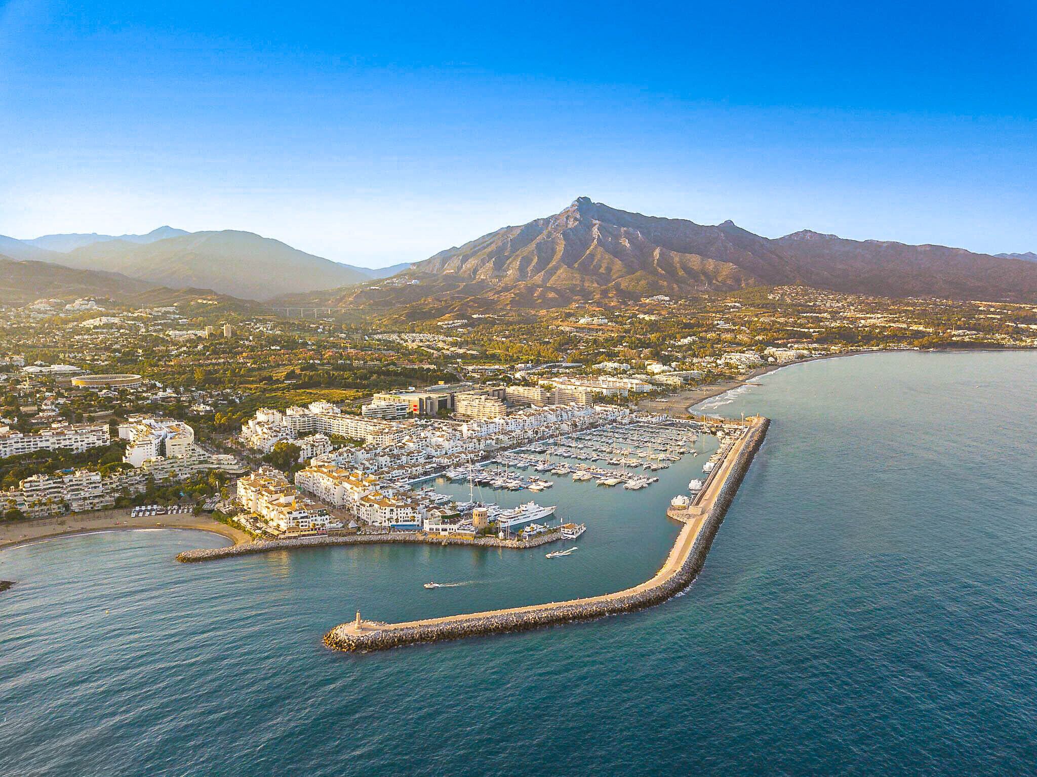 Vista aèria d'una població costanera amb un port esportiu, edificis residencials i una muntanya destacada al fons.