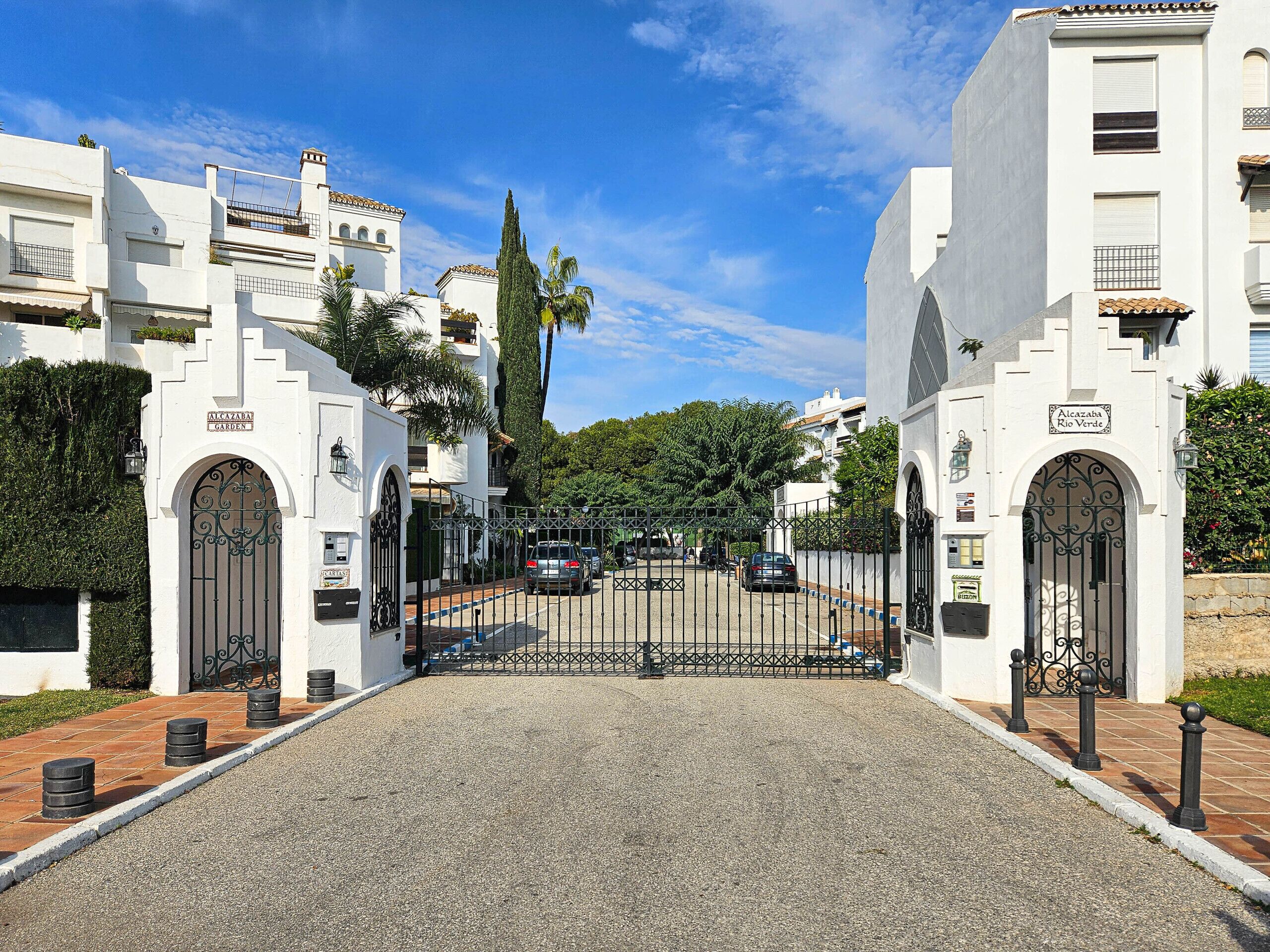 A gated entrance to a residential area with white buildings, a clear sky, and a gravel driveway.