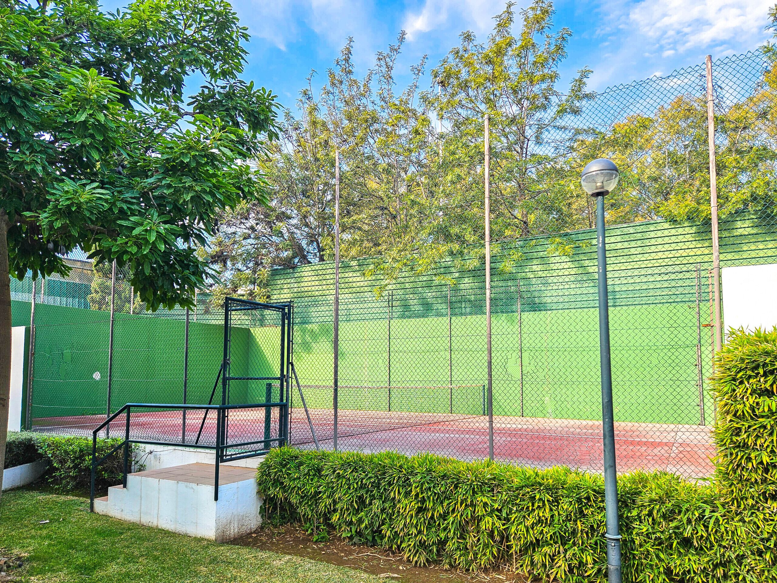 A fenced tennis court with a metal frame and steps leading up to it, surrounded by greenery and a clear sky.