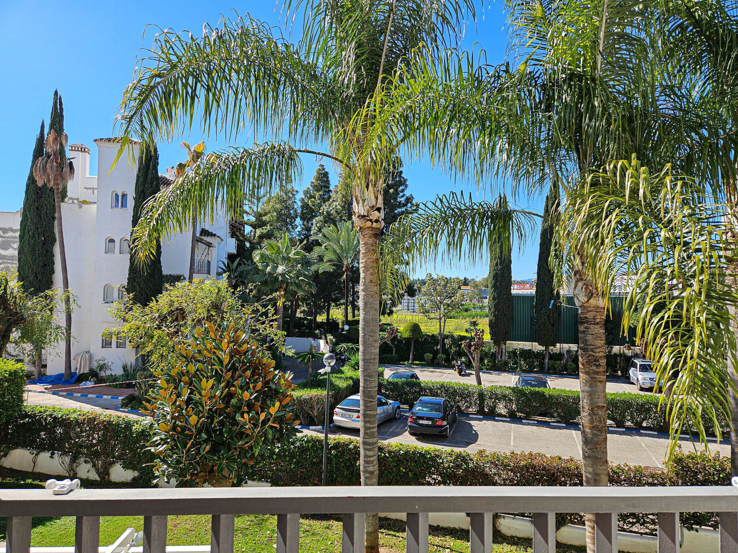 A residential area with a white building, palm trees, and parked cars.