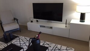 Ein Wohnzimmer mit Flachbildfernseher auf einer weißen Unterhaltungsanlage, einem schwarzen Couchtisch mit Fernbedienung, einem gemusterten Teppich und einem Sessel.