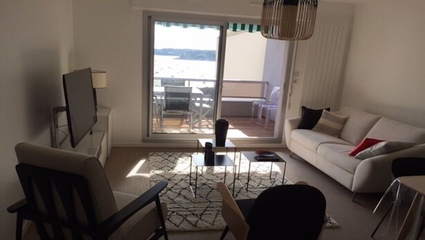 Ein modernes Wohnzimmer mit einer Couch, Sesseln, einem Fernseher und einem Balkon mit Gartenmöbeln.