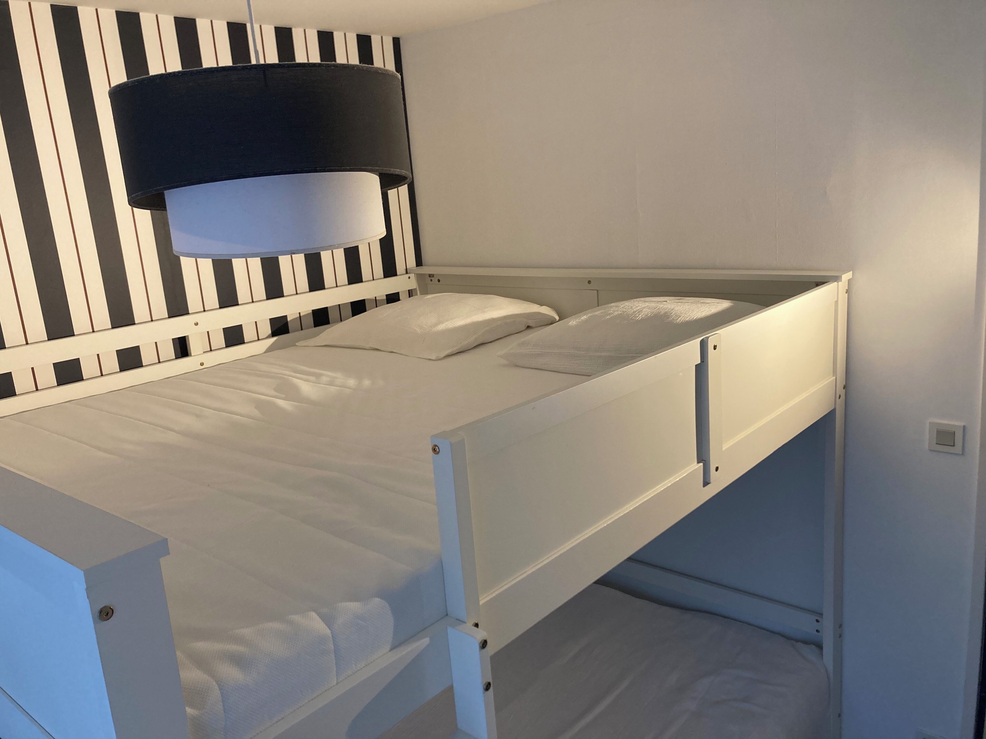 En køjeseng med hvid madras og puder, væg med sorte og hvide striber og en sort lampe over sengen.