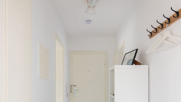 Een smalle gang met een houten vloer, een witte deur aan het einde en een wit schap met twee zwarte kussens en een ingelijst beeldje erop.