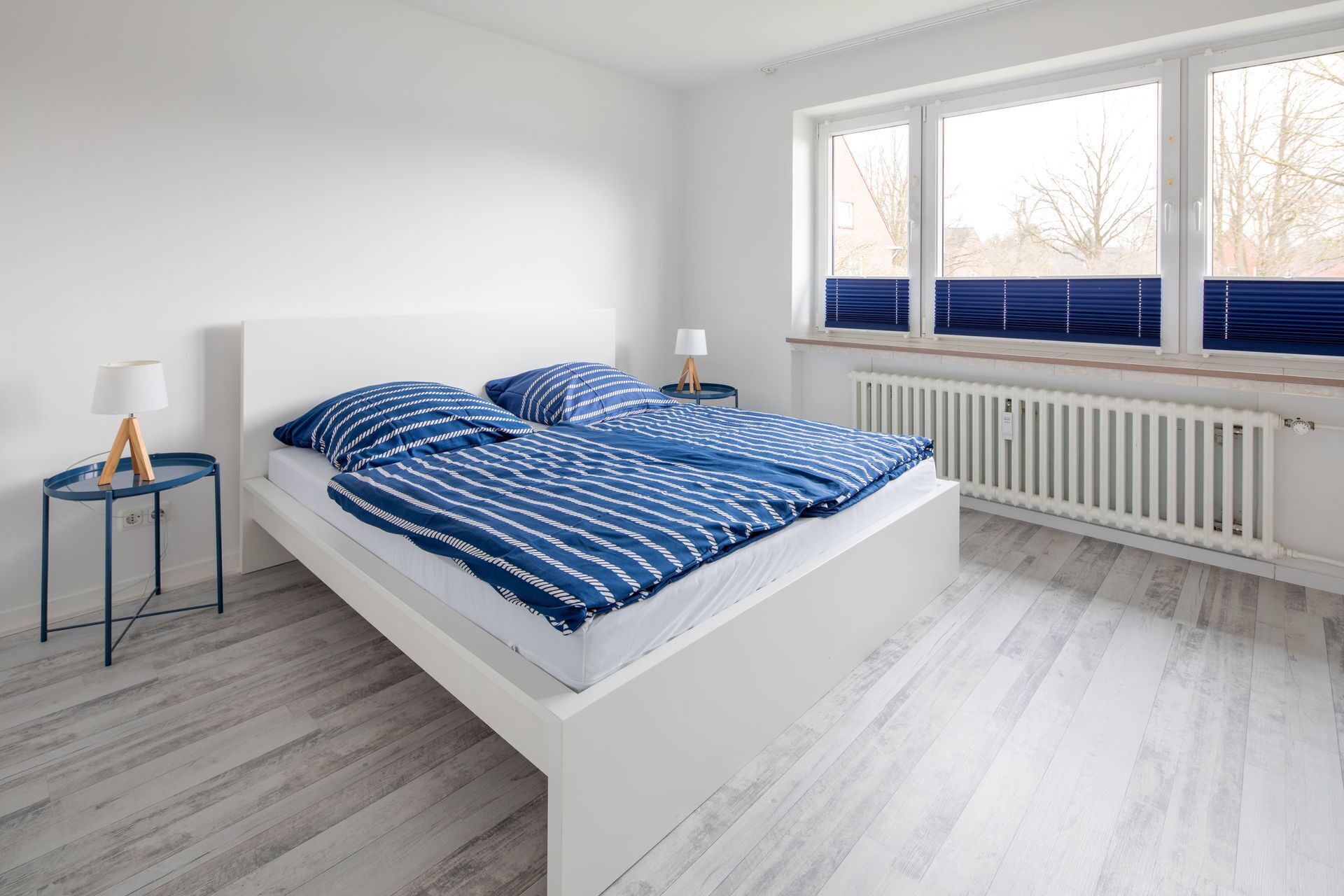 Ein modernes Schlafzimmer mit einem weißen Bett, blau gestreifter Bettwäsche, zwei Nachttischen mit Lampen und einem großen Fenster mit Jalousien.