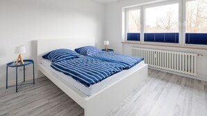 Een moderne slaapkamer met een wit bed, beddengoed met blauwe strepen, twee nachtkastjes met lampen en een groot raam met jaloezieën.