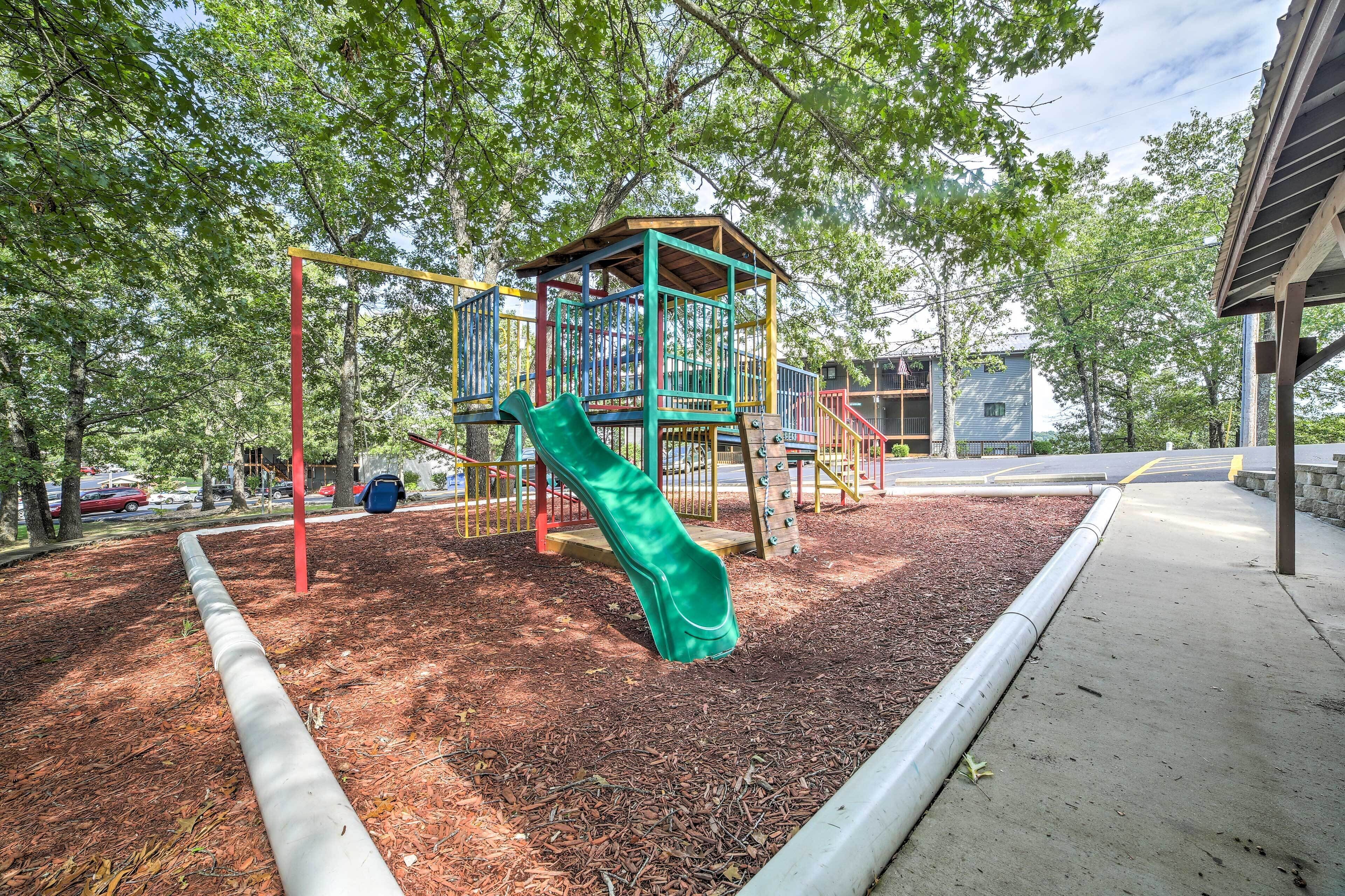 A playground with a slide and climbing structures, surrounded by trees and a paved walkway.