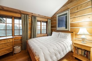 A wooden cabin bedroom with a large bed, a nightstand with a lamp, a bookshelf, and a window with curtains. - Canyon Creek Ranch - Wonderful home located in the Tom Miner Basin! (Emigrant)