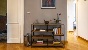 Un pasillo con suelo de madera, una puerta blanca, una estantería con libros, un cuadro en la pared y una pequeña estatua en el suelo.