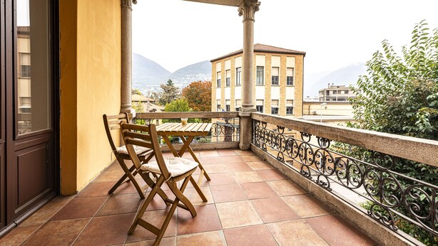 Un balcón con mesa y sillas, con vista a un paisaje urbano y montañas al fondo.