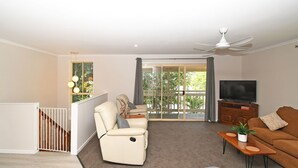 Ein Wohnzimmer mit einer Couch, einem Fernseher, einem Deckenventilator und einer Schiebeglastür, die auf einen Balkon führt.