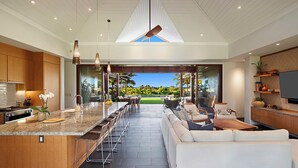 A modern kitchen with a central island, a dining area with chairs, a white sofa, and a TV on a wooden shelf. - Hale Kai Malia: Spacious, Stylish Ocean View Villa (Koloa)