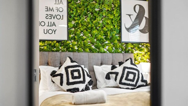 A neatly made bed with geometric patterned pillows, a wall with green foliage, and two framed signs with text.