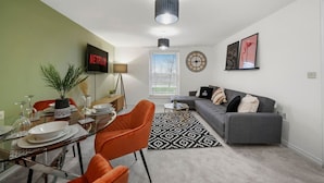 A modern living room with a glass dining table, orange chairs, a grey sofa, and a black and white patterned rug.