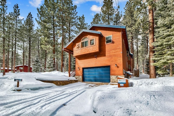 Une cabane en bois à deux étages, avec une porte de garage bleue, entourée de grands pins et d’un sol recouvert de neige.