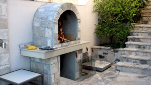 Een buitenoven van steen met een vuur erin, een metalen grill op het aanrecht, en een tafel met een metalen frame.