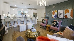 Ein modernes Wohnzimmer mit einer Couch, einem Hocker und einem Couchtisch, eine Küche mit weißen Schränken und einem Kühlschrank sowie ein Essbereich mit Stühlen und einem Tisch.