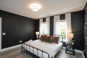 A bedroom with a black and white color scheme, a bed with multiple pillows, a bedside table with a lamp, and a window with curtains. - 🥳​🎉24 Beds.  Large CELEBRATION Home.  5 Points Section.  Close to the Heart of T (Nashville)