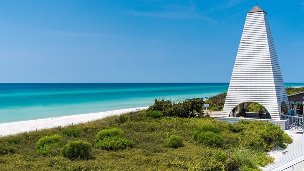 Ett högt, vitt, pyramidformat monument med en spetsig topp, som står på en strand med kristallklart turkosfärgat vatten och vit sand.