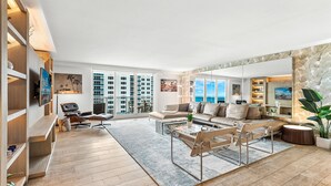 A spacious living room with a large window offering a view of high-rise buildings and the sea. There is a comfortable sofa set, a coffee table, and a bookshelf.